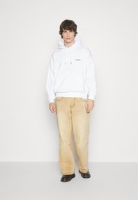 Felpa bianca in cotone con cappuccio e cordino, caratterizzata da un piccolo logo applicato, abbinata a jeans a gamba larga beige e scarpe nere.