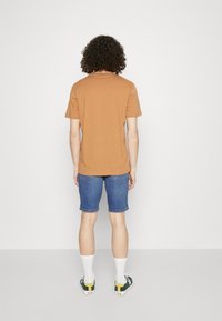 Camiseta de algodón marrón, de mangas cortas, vista trasera. Llevada con pantalones cortos de mezclilla azules, calcetines blancos y zapatillas verdes con detalles amarillos.
