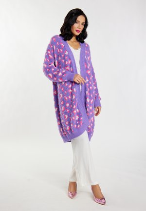 Paarse fluffy cardigan met een abstract patroon in roze en wit, losse pasvorm, open voorkant, gecombineerd met witte broek en glanzende roze hakken.