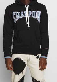 Felpa nera con cappuccio e cordino bianco, logo "CHAMPION" ricamato in azzurro. Presenta una tasca a canguro e polsini con un accento blu.