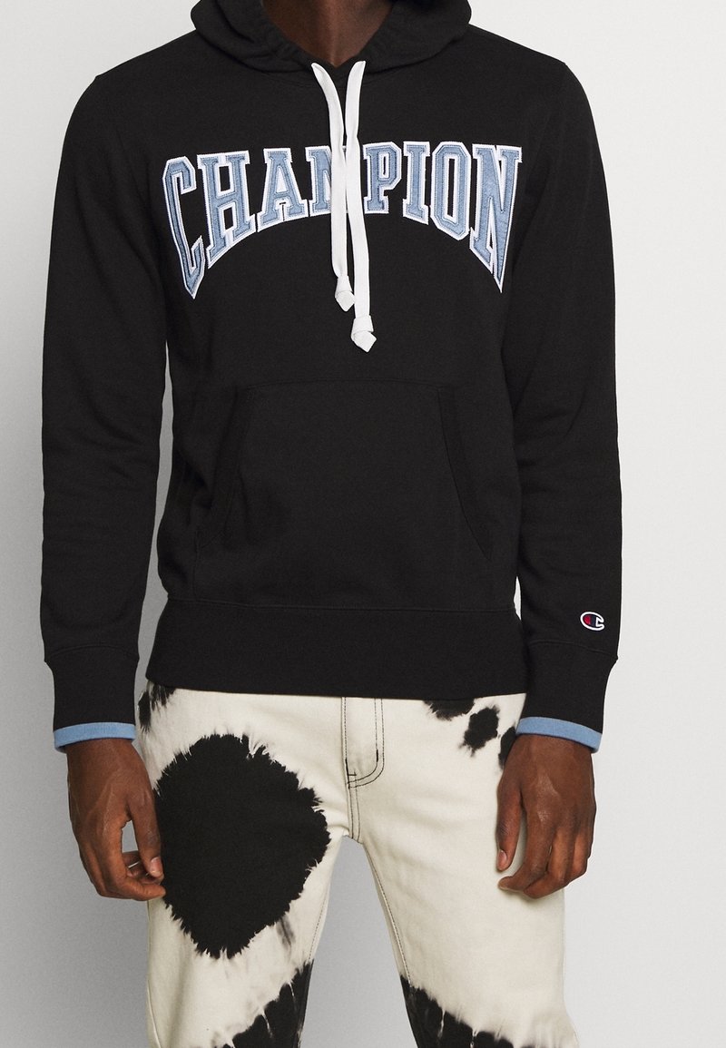 Felpa nera con cappuccio e cordino bianco, logo "CHAMPION" ricamato in azzurro. Presenta una tasca a canguro e polsini con un accento blu.