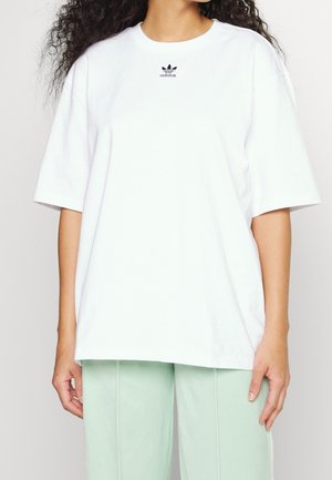 Kvinde iført en oversized hvid Adidas T-shirt med logo på brystet, kombineret med lysegrønne bukser.