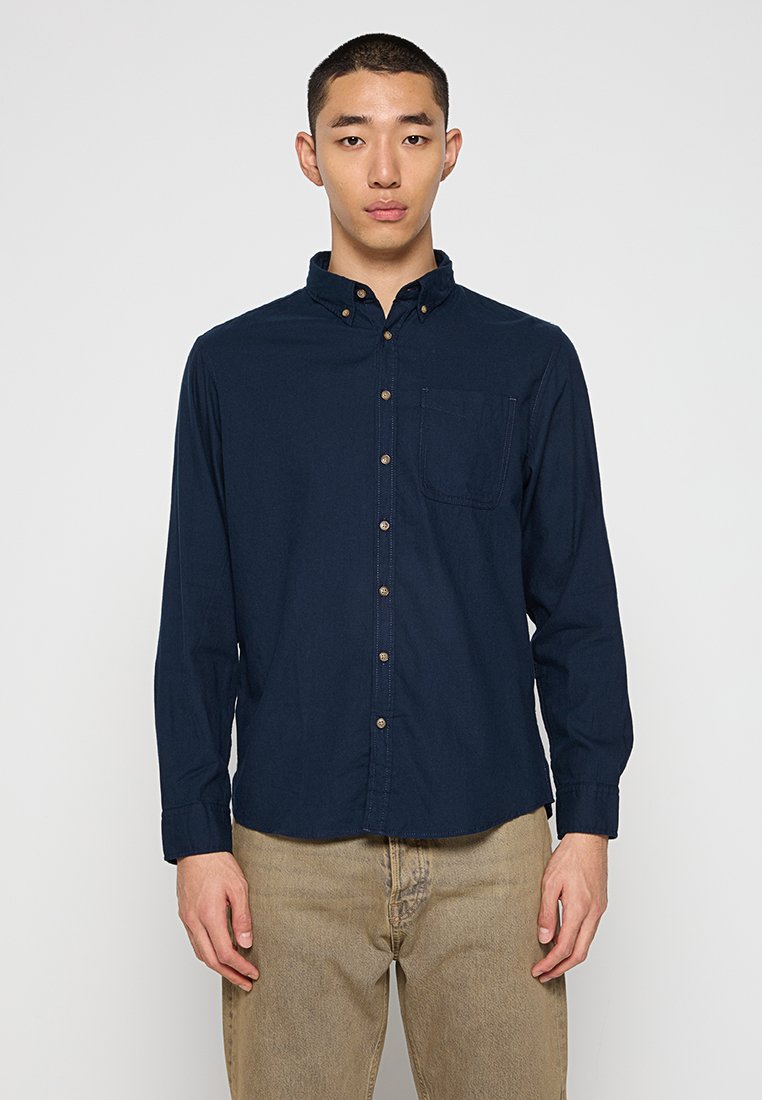 Camisa azul marino de botones con un cuello puntiagudo, mangas largas, un bolsillo en el pecho y botones marrones; hecha de un material de algodón suave.