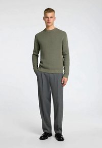 Camisola de malha verde-oliva com decote canelado, combinada com calças cinzentas plissadas e sapatos pretos. Texturas suaves e silhuetas ajustadas.