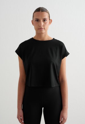 Vrouw met naar achteren gekamd haar die een losse zwarte cropped t-shirt en nauwsluitende zwarte broek draagt, staand tegen een effen lichte achtergrond.