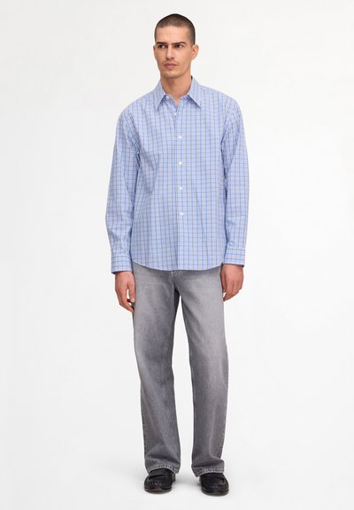 Homme debout portant une chemise à carreaux bleue à manches longues, un jean gris clair et des chaussures noires sur un fond blanc uni.