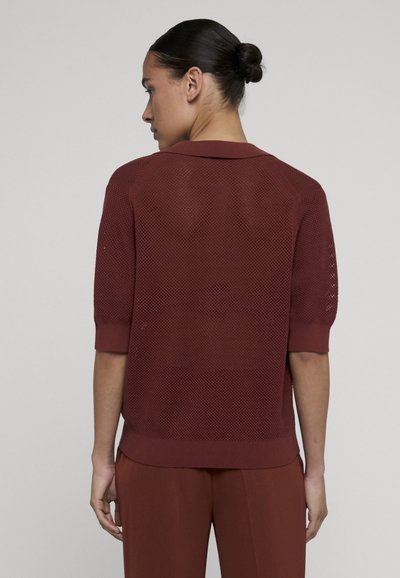 Femme aux cheveux relevés en chignon, vêtue d'un haut en maille bordeaux à manches courtes et d'un pantalon assorti, dos au spectateur sur un fond uni.