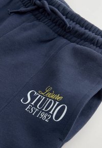 Pantalons de jogging navy en tissu doux, dotés d'une taille élastique, d'un cordon de serrage et d'un texte imprimé blanc et doré : "Leisure STUDIO EST 1982."