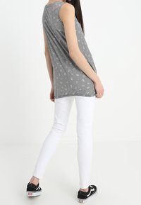 Kvinna i en utanärmad svart och vit randig topp, vita skinny jeans och svarta sneakers, stående mot en enkel vit bakgrund.