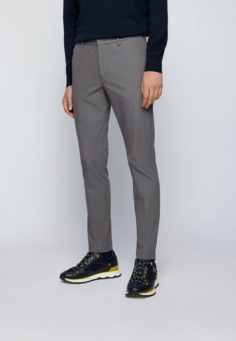 Homme portant un pantalon gris slim, des baskets noires et jaunes, et un haut à manches longues noir devant un fond uni.
