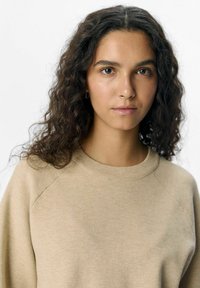Beige trui met een ronde hals en raglanmouwen, met een zachte, gestructureerde brei. Model heeft golvend bruin haar en minimale accessoires.