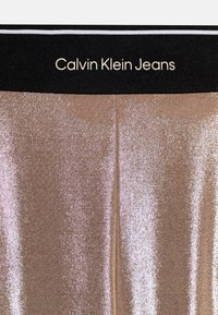 Metalliske gullleggings med en skinnende tekstur, med et svart liv som har påskriften "Calvin Klein Jeans" i gullbokstaver.