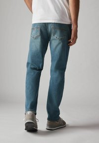 Jeans de mezclilla azul con corte recto, con costuras naranjas y dos bolsillos traseros, combinados con zapatillas grises.