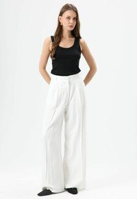 Débardeur noir côtelé associé à un pantalon blanc à jambes larges, présentant une taille haute, des plis subtils et une silhouette décontractée. Chaussures pointues noires.