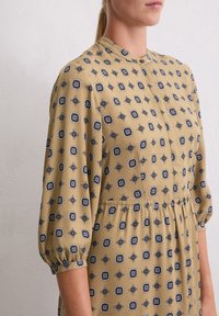 Langarm, beige jurk met zwarte geometrische patronen, een ronde kraag, knoopsluiting en elastische taille, gemaakt van gladde stof.
