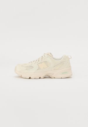 Beige low-top athletic sneaker with mesh panels, lace-up front, cushioned sole, and branding on the side and heel.