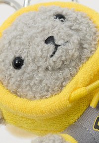 Αφράτος γκρι αρκούδος με σγουρό τρίχωμα, ντυμένος με ένα κίτρινο φούτερ με κουκούλα και κρατώντας ένα γκρι αντικείμενο με κίτρινο κορδόνι.