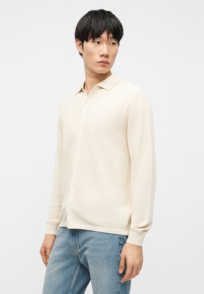 Jeune homme aux cheveux noirs portant une chemise crème à manches longues boutonnée et un jean bleu clair, regardant sur le côté contre un fond uni.