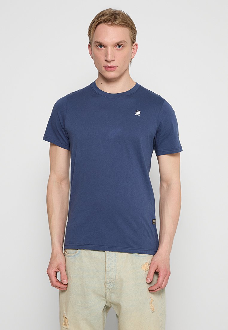 Marineblaue Baumwoll-T-Shirt mit rundem Ausschnitt, kurzen Ärmeln und kleinem Logo auf der linken Brust. Getragen mit hellen, stark verwaschenen Jeans.