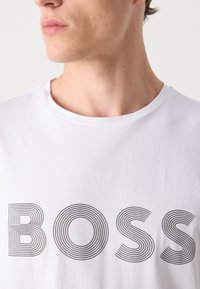 Witte katoenen T-shirt met het woord "BOSS" in vetgedrukte, golvende zwarte lijnen. Het heeft een ronde hals en korte mouwen.