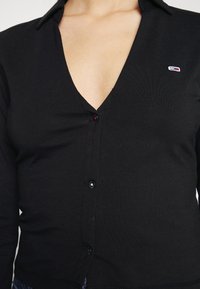 Black cardigan with a V-neck design, featuring a button closure and small logo accent. Smooth texture and ribbed fabric detail.