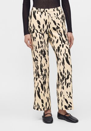 Femme portant un pantalon crème droit avec des taches abstraites noires, associé à un top noir et des chaussures plates noires à brides.