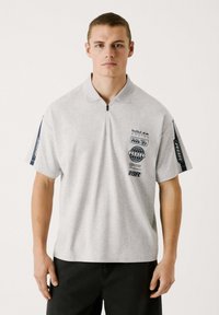Siva polo majica s kratkimi rokavi, zadrgo na ovratniku in črnimi detajli. Ima tiskane logotipe in grafike na sprednjem delu in rokavih.