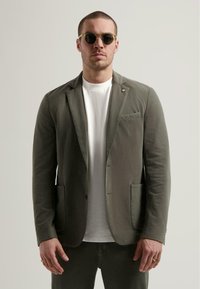 Oliwkowa bawełniana marynarka z wyłogami, dwiema przednimi kieszeniami i małą kieszonką na piersi. Nosi się ją na białym t-shircie. Okrągłe okulary przeciwsłoneczne.