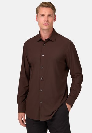 Chemise marron à manches longues avec un col classique, dotée d'une fermeture à boutons et d'un tissu lisse. Associée à un pantalon sombre, soulignant une apparence soignée.