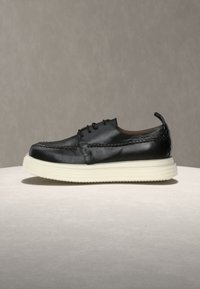 Μαύρο καθημερινό δερμάτινο παπούτσι με παχιά λευκή καουτσούκ σόλα και κορδόνια μπροστά, τοποθετημένο σε ανοιχτό γκρι επιφάνεια και ουδέτερο φόντο.