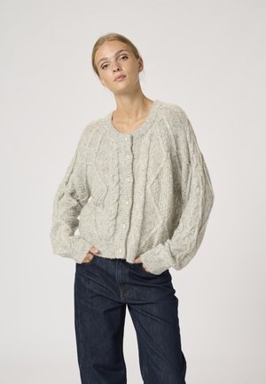 Cardigan en tricot gris clair avec motif en câble et boutons en perles, présentant des épaules tombantes et des poignets côtelés, porté sur un jean foncé.