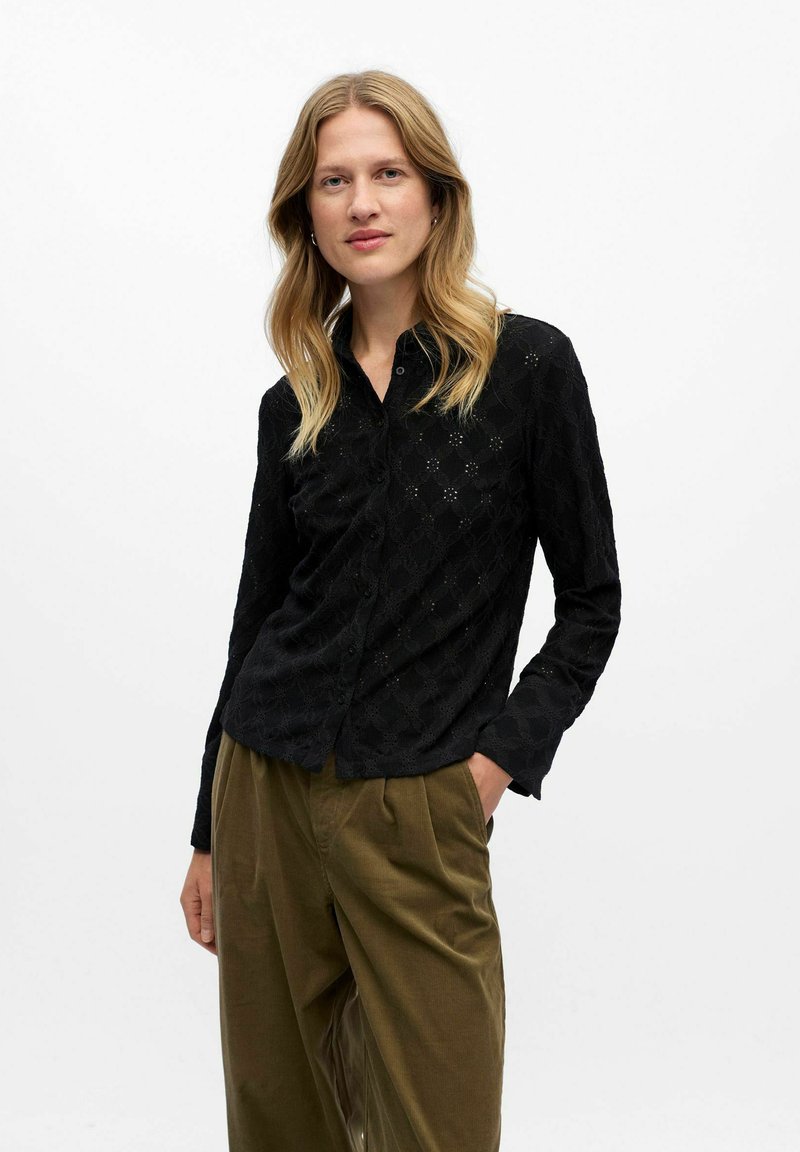 Chemise noire texturée à manches longues avec un motif floral, associée à un pantalon plissé vert olive.