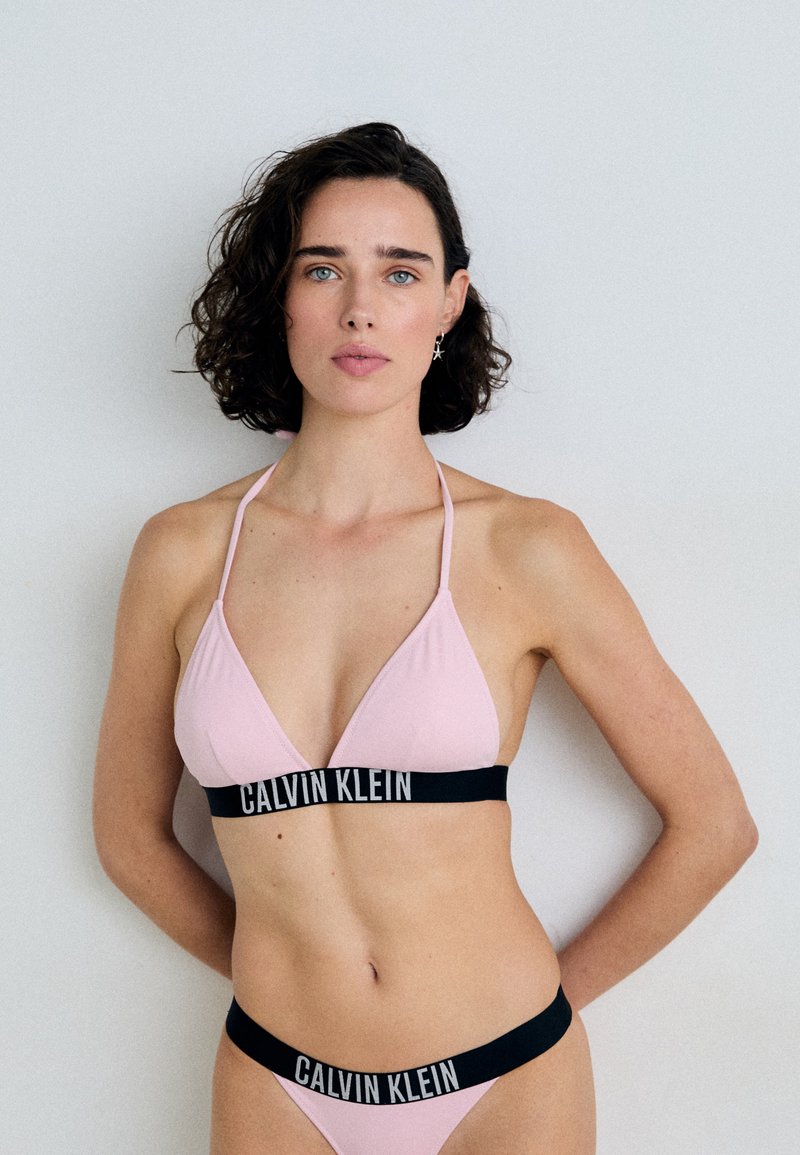 Pinkfarbener, dreieckiger Bikini-Oberteil mit schlanken Trägern und schwarzem Bund, der das „CALVIN KLEIN“-Logo trägt; passender Bikini-Unterteil mit einem ähnlichen Bunchendesign.