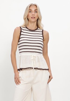 Vrouw met lang blond haar draagt een mouwloze, bruin-wit gestreepte gebreide top met peplumzoom en crèmekleurige broek, staand tegen een effen achtergrond.