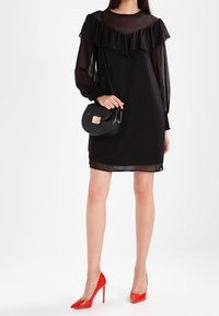 Femme portant une robe noire transparente à manches longues avec un détail à volants, tenant un sac à main noir et portant des chaussures à talons rouges.