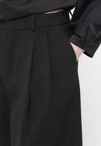 Schwarze, plissierte Hosen mit tailliertem Bund und seitlichen Taschen, aus einem glatten, strukturierten Stoff gefertigt.