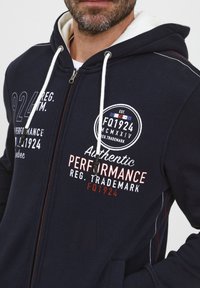 Felpa con zip di color navy, con fodera interna bianca, caratterizzata da testo stampato e accenti di logo in bianco e rosso. Cords con punte nere.