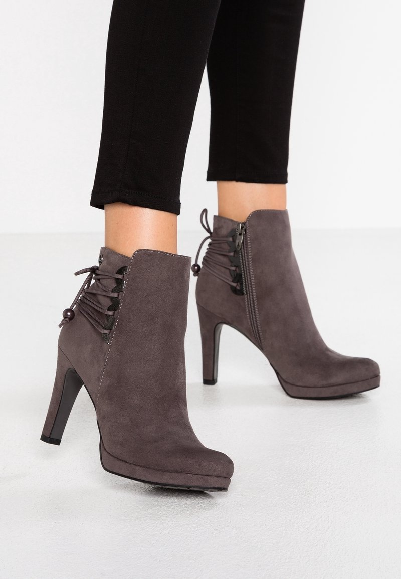 Femme portant des bottines en suède gris foncé à talons hauts, fermeture éclair latérale et détail décoratif à lacets à l'arrière, assorties à un pantalon noir.