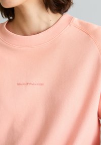 Nahaufnahme einer Person, die ein hellrosa Sweatshirt mit der Prägung "Marc O'Polo DENIM" auf der Brust trägt.