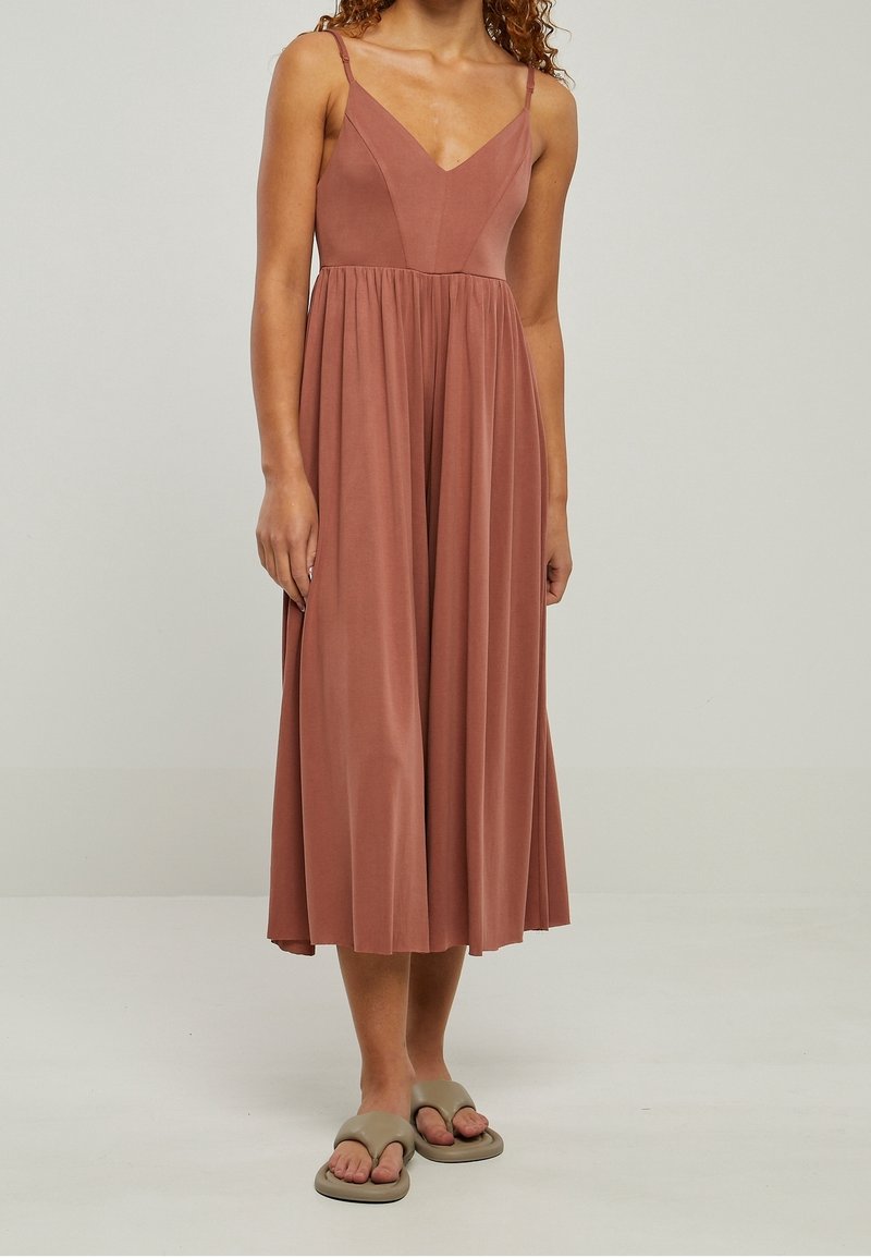 Femme portant une robe midi rose poudré sans manches avec des bretelles fines et une jupe plissée, associée à des sandales beiges à bout ouvert sur un fond clair.