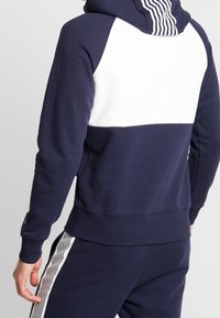 Sudadera azul marino con una franja blanca horizontal en la parte posterior de los hombros. Presenta puños y cinturilla acanalados, con acentos de rayas verticales en la capucha.