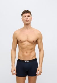 Μαύρα αντρικά μποξέρ με το λογότυπο "BOSS" στη ζώνη, φορεμένα από μοντέλο με γυμνασμένο σώμα, που στέκεται σε ουδέτερο περιβάλλον.