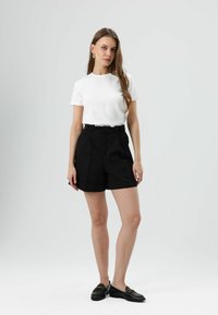 T-shirt blanc à col rond associé à un short noir taille haute, doté de plis et d'une coupe ajustée, complété par des mocassins noirs.