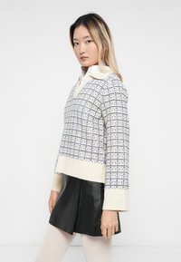 Jonge vrouw met lang haar, gekleed in een gebreide trui met patroon, witte blouse, zwarte leren rok en witte panty, tegen een effen achtergrond.