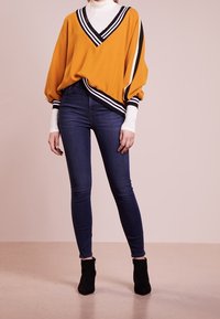 Suéter oversize mostaza con cuello en V a rayas en blanco y negro, combinado con jeans ajustados azul oscuro y botas de tobillo negras. Textura suave.