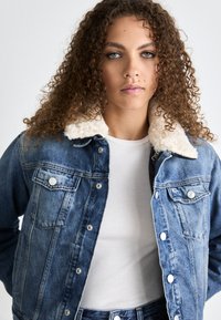Chaqueta de mezclilla con un lavado azul claro, cuello de sherpa, cierres de botones y dos bolsillos en el pecho. Llevada sobre una camisa blanca.
