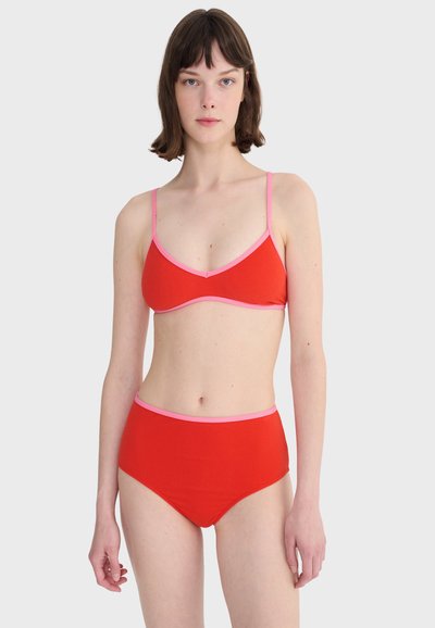 Femme aux cheveux courts et foncés, portant un bikini rouge avec des bordures roses, composé d'un haut triangle et d'un bas taille haute, debout devant un fond uni.