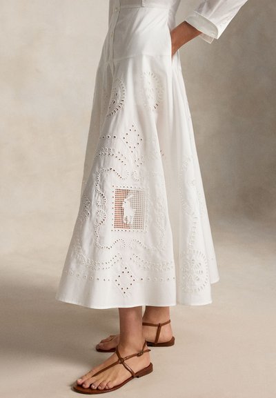 Femeie purtând o fustă midi albă din material brodat cu buzunare și sandale maro din piele cu bretele, stând în fața unui fundal neutru.