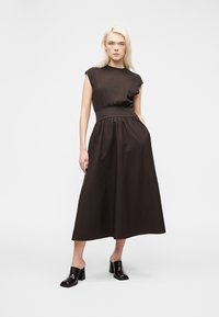 Blonde vrouw staand in een mouwloze zwarte midi-jurk met getailleerde taille en zwarte hakmuiltjes op een witte achtergrond.