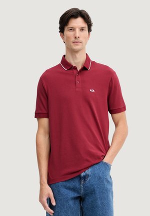 Uomo che indossa una polo color amaranto con rifiniture bianche e logo, abbinata a jeans blu, in piedi davanti a uno sfondo chiaro uniforme.